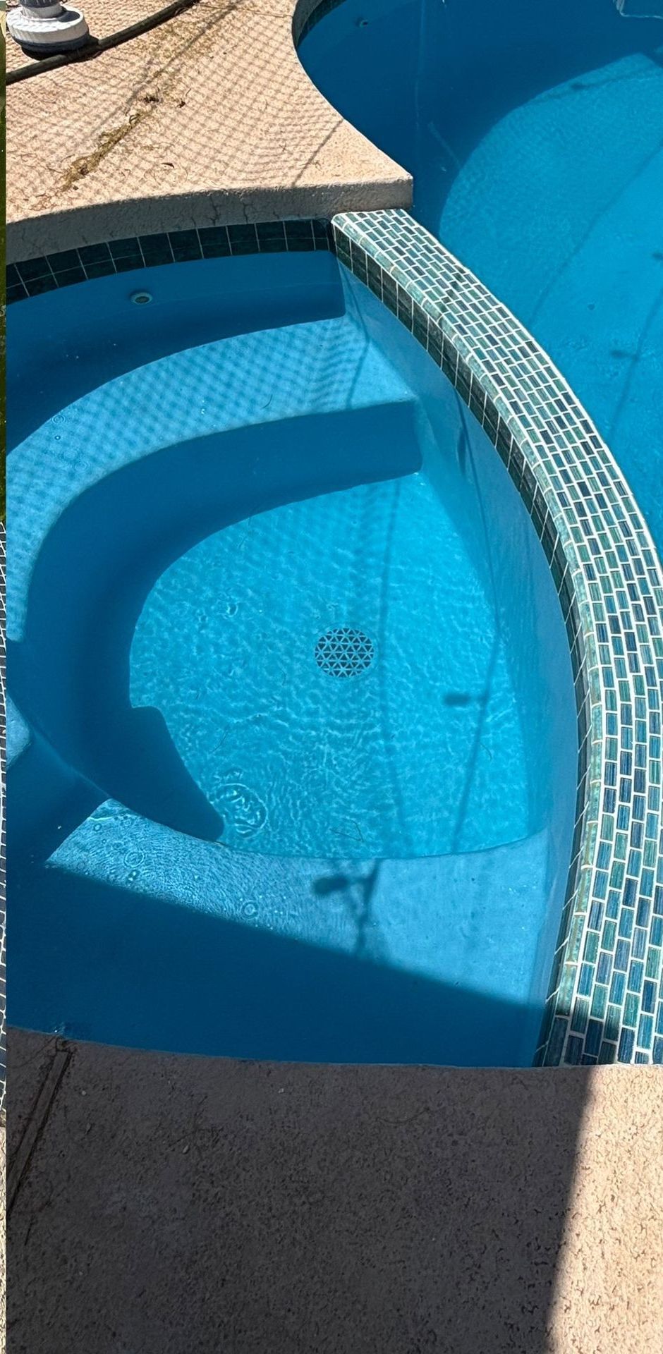 A high-angle view of pool steps submerged in clear blue water, adjacent to a decorative tiled edge and a concrete deck.