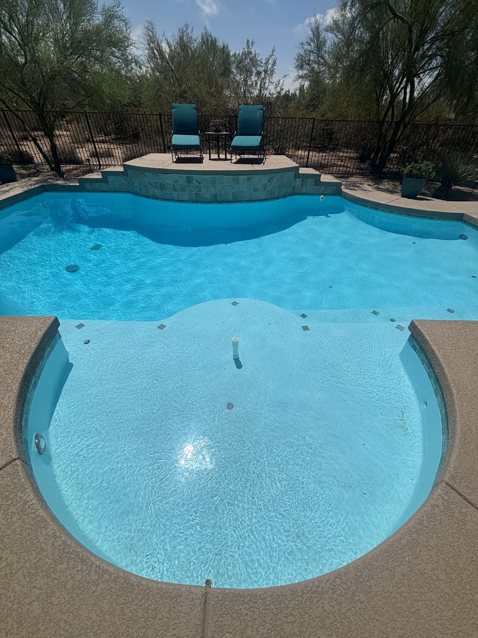 Blue swimming pool with shallow spa area and two lounge chairs in the background under trees
