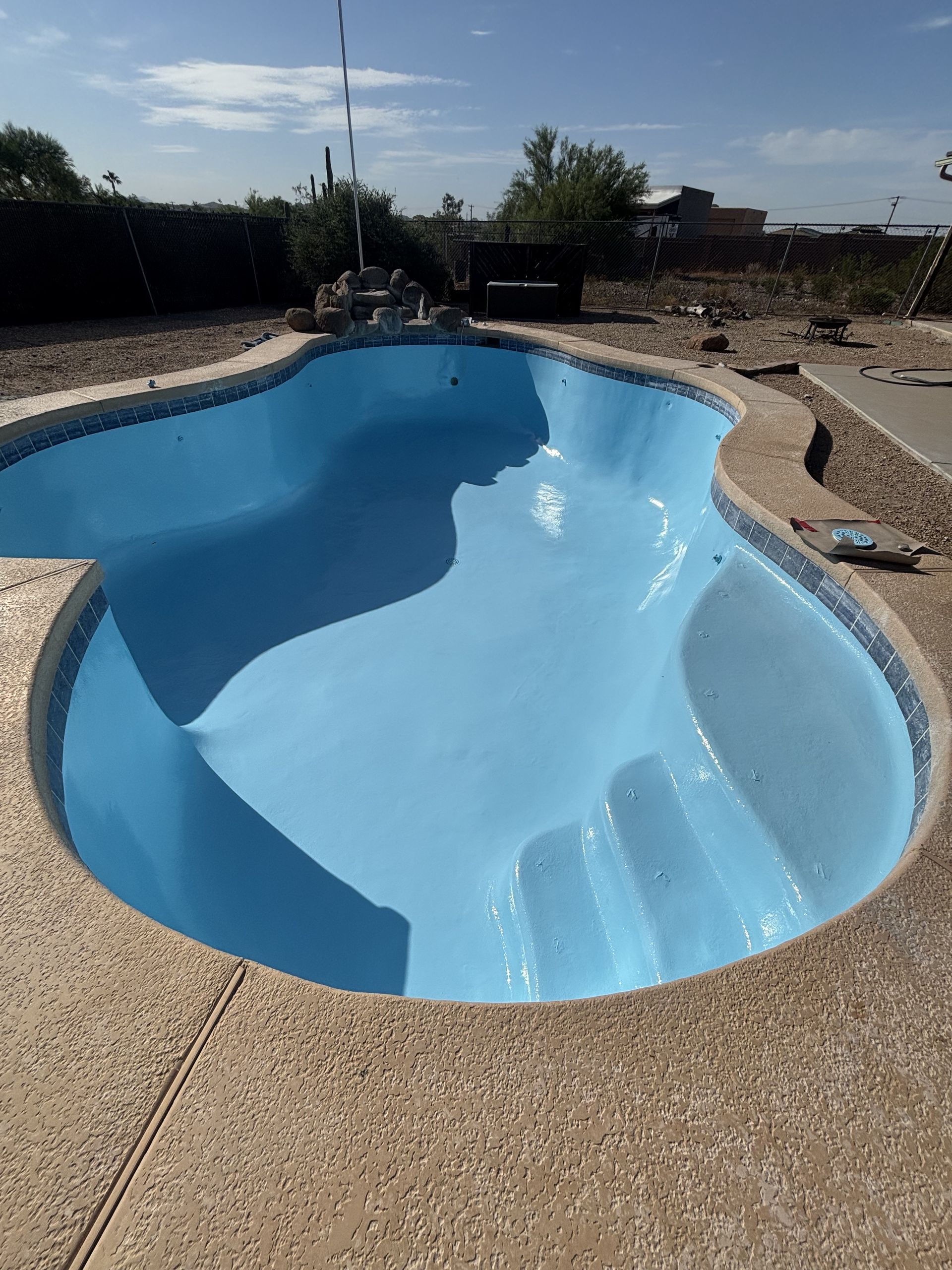 Empty blue backyard swimming pool with a curved shallow end and stone deck under a clear sky