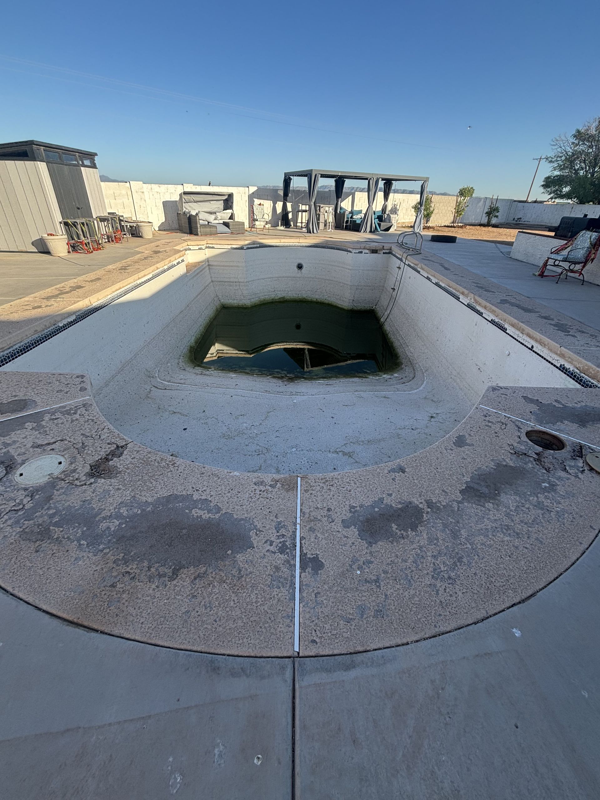 Empty outdoor pool with a large hole in the deep end, surrounded by concrete under clear blue sky