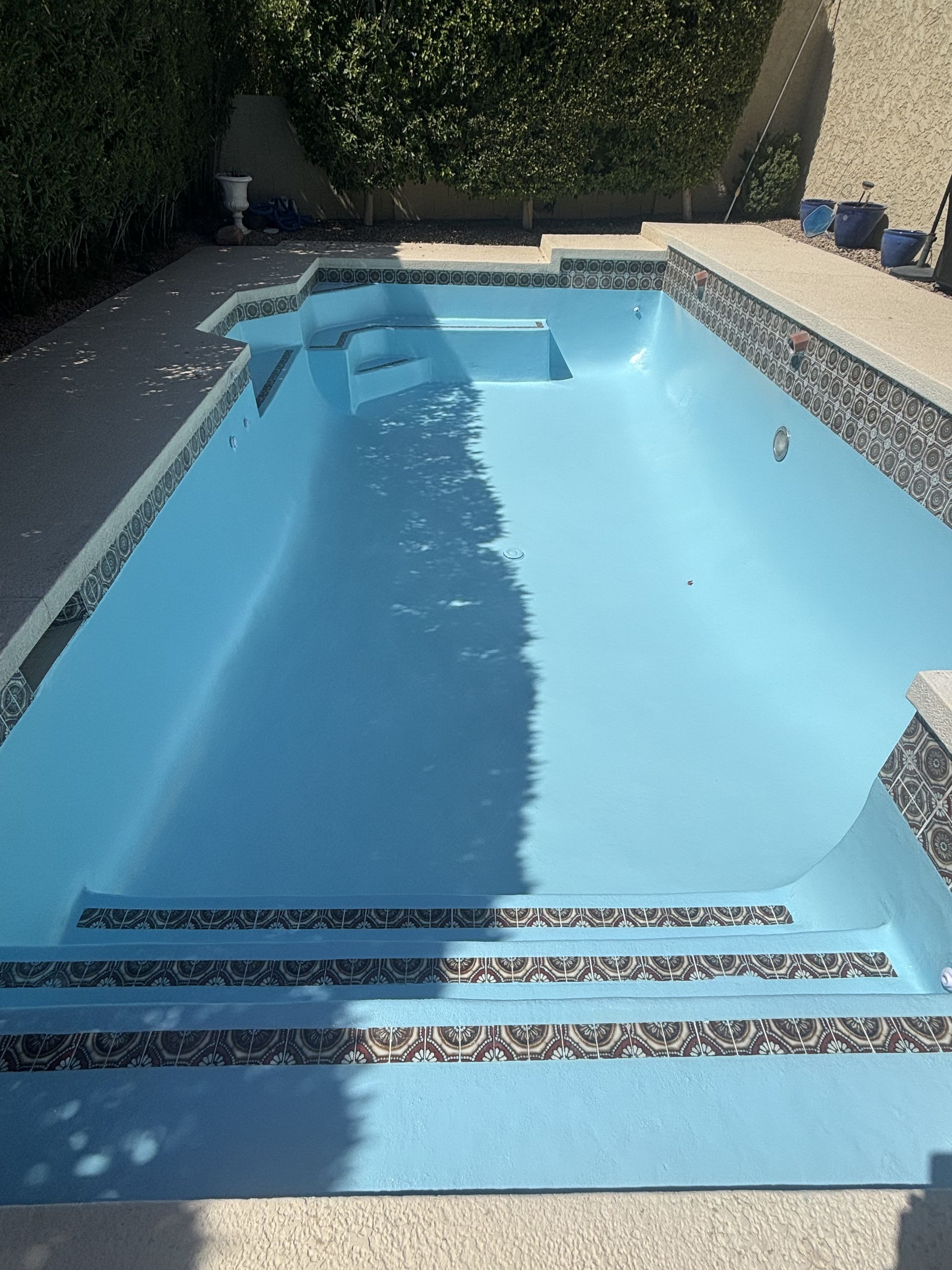 Outdoor swimming pool with blue water, steps, and a shaded patio beside a hedge-lined wall