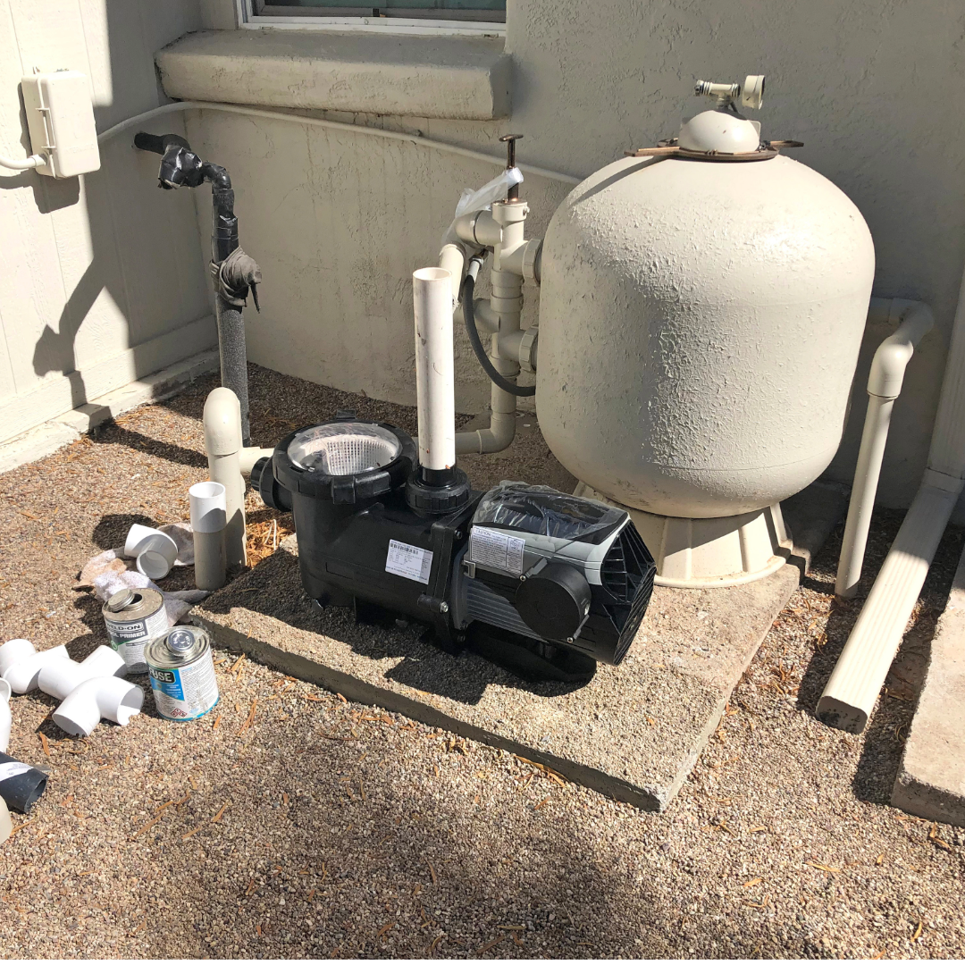 A black pool pump connected to white PVC pipes and a beige sand filter on a concrete pad against a beige wall.