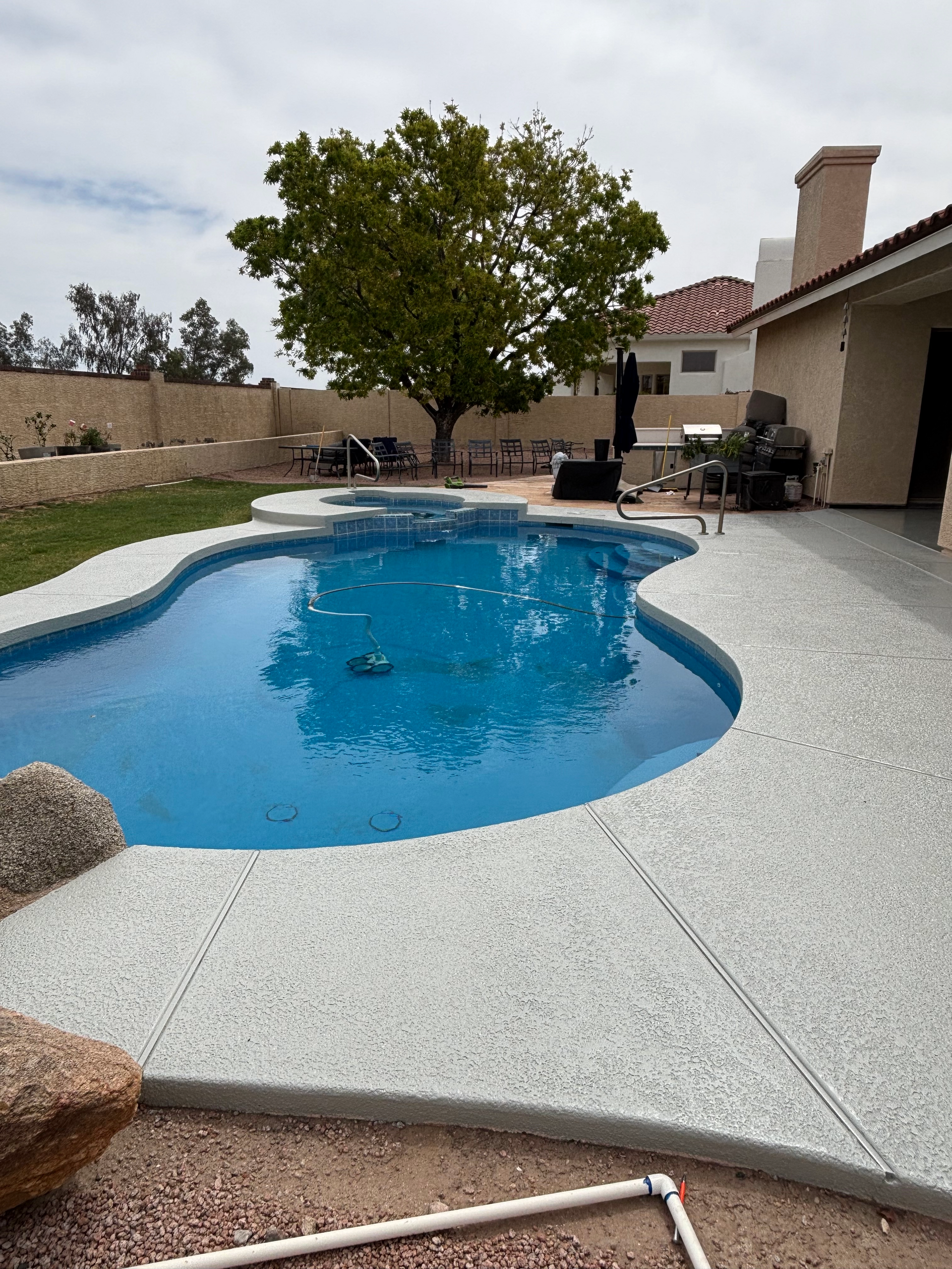 A kidney-shaped swimming pool with a spa and light grey textured decking in a residential backyard.