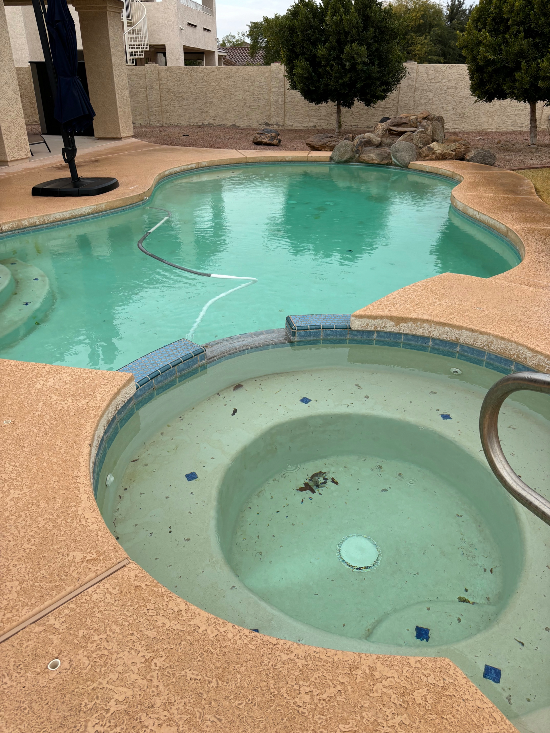 A backyard scene featuring a round spa in the foreground next to a larger swimming pool with light-colored decking.