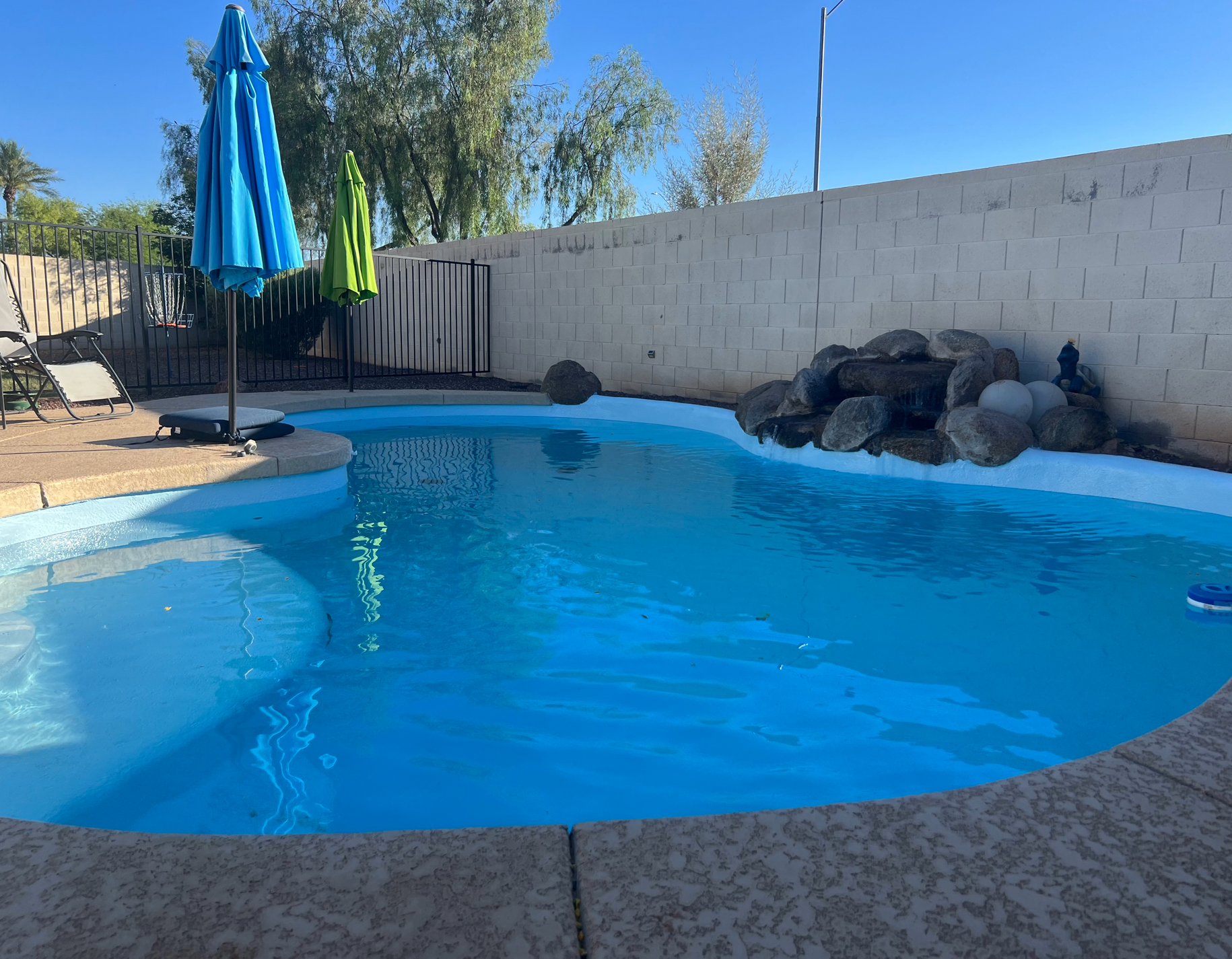 A backyard swimming pool with a rock water feature, a tan deck, and blue and green patio umbrellas on a sunny day.