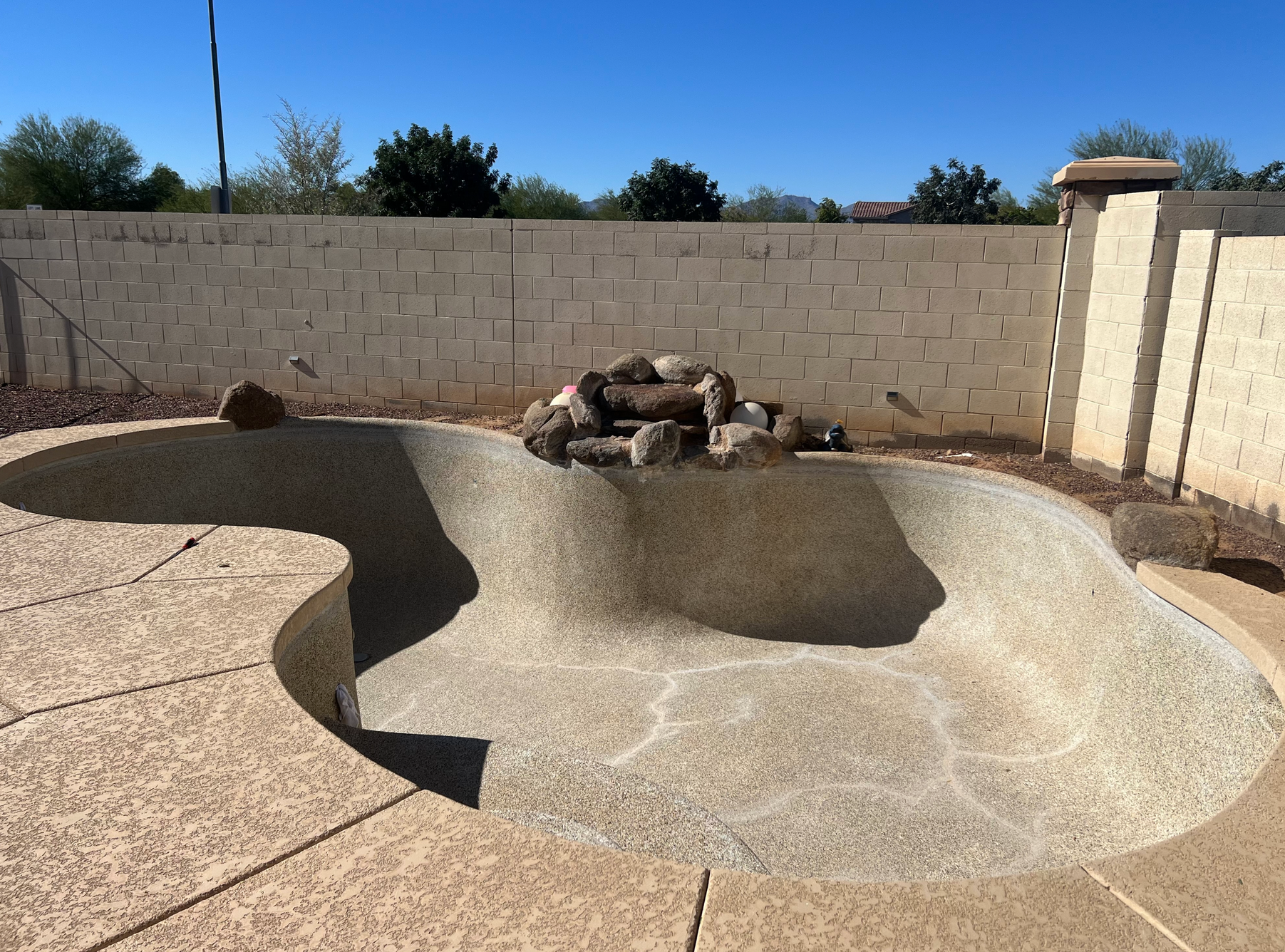An empty, textured-finish swimming pool with a stone rock feature in the center, set against a tan cinder block wall.