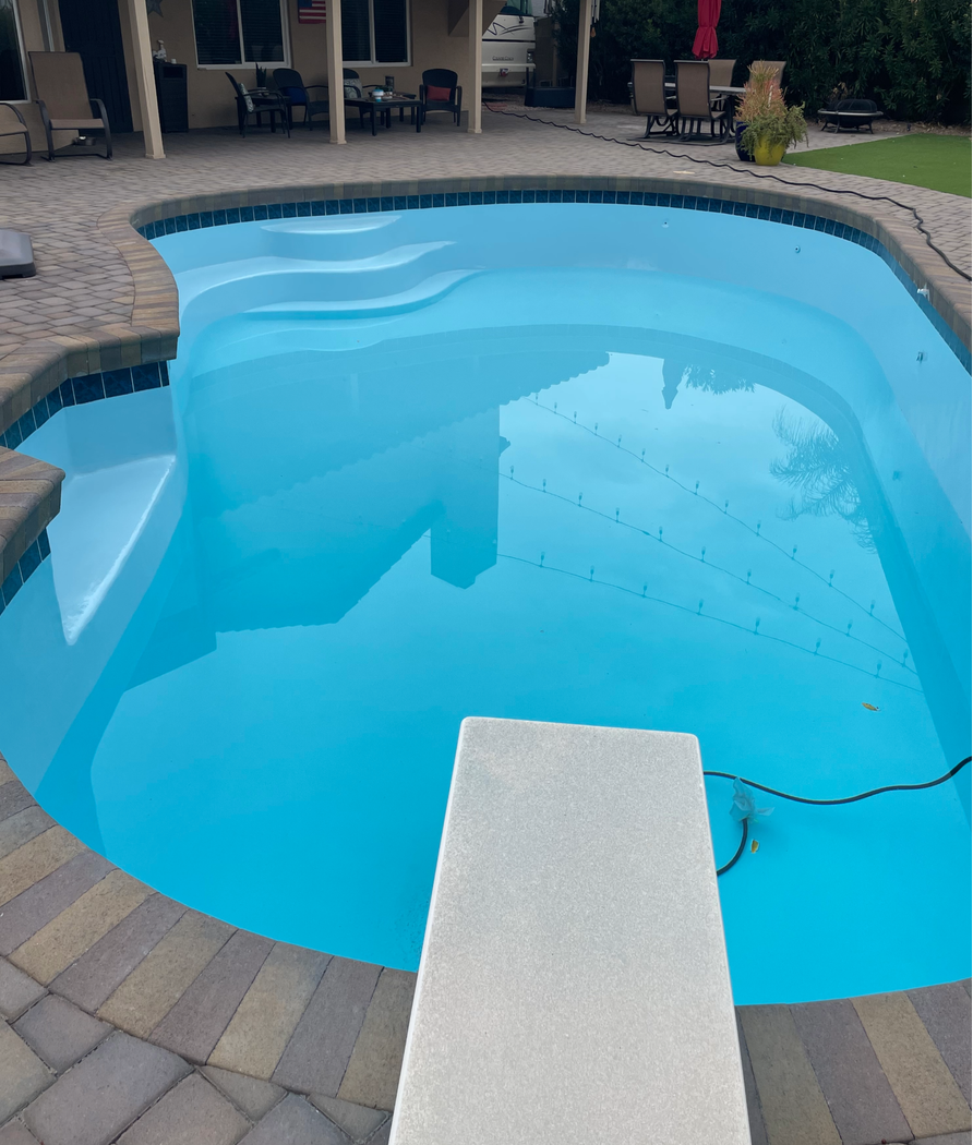 A backyard swimming pool with light blue water and a diving board extending from the stone patio edge.