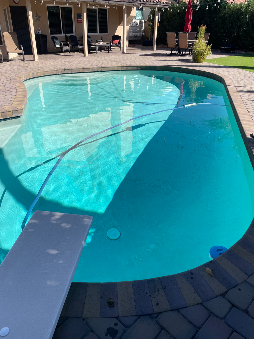 A residential backyard swimming pool with a diving board, surrounded by a paved patio area.