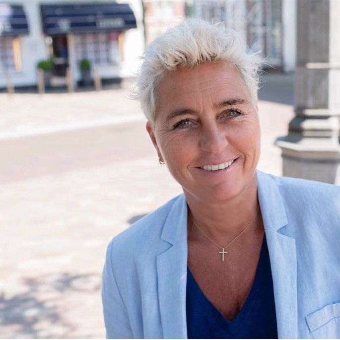 Een persoon met kort, lichtgekleurd haar en een lichtblauwe blazer lacht buiten voor een gebouw.