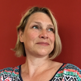 Een persoon met blond haar en een overhemd met een patroon kijkt weg van de camera, tegen een effen rode achtergrond.