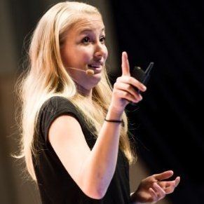 Een vrouw in een zwart shirt spreekt op het podium, terwijl ze een zendapparaat vasthoudt en met een opgestoken wijsvinger gebaart.
