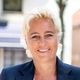 Een persoon met kort blond haar glimlacht hartelijk terwijl ze een donkere blazer en een klein kruisje om haar nek draagt.