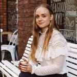 Een persoon met lang blond haar, gekleed in een witte top met kanten details, zit buiten op een witte bank met een kop koffie in haar hand.