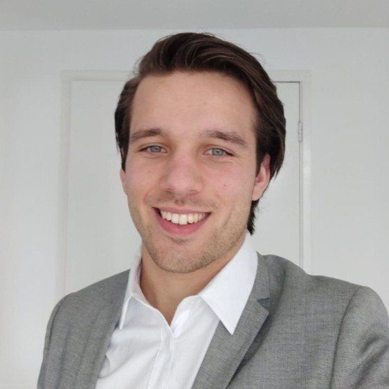 Een persoon met donker haar, gekleed in een grijs colbert en een wit overhemd, glimlacht voor een effen witte muur.