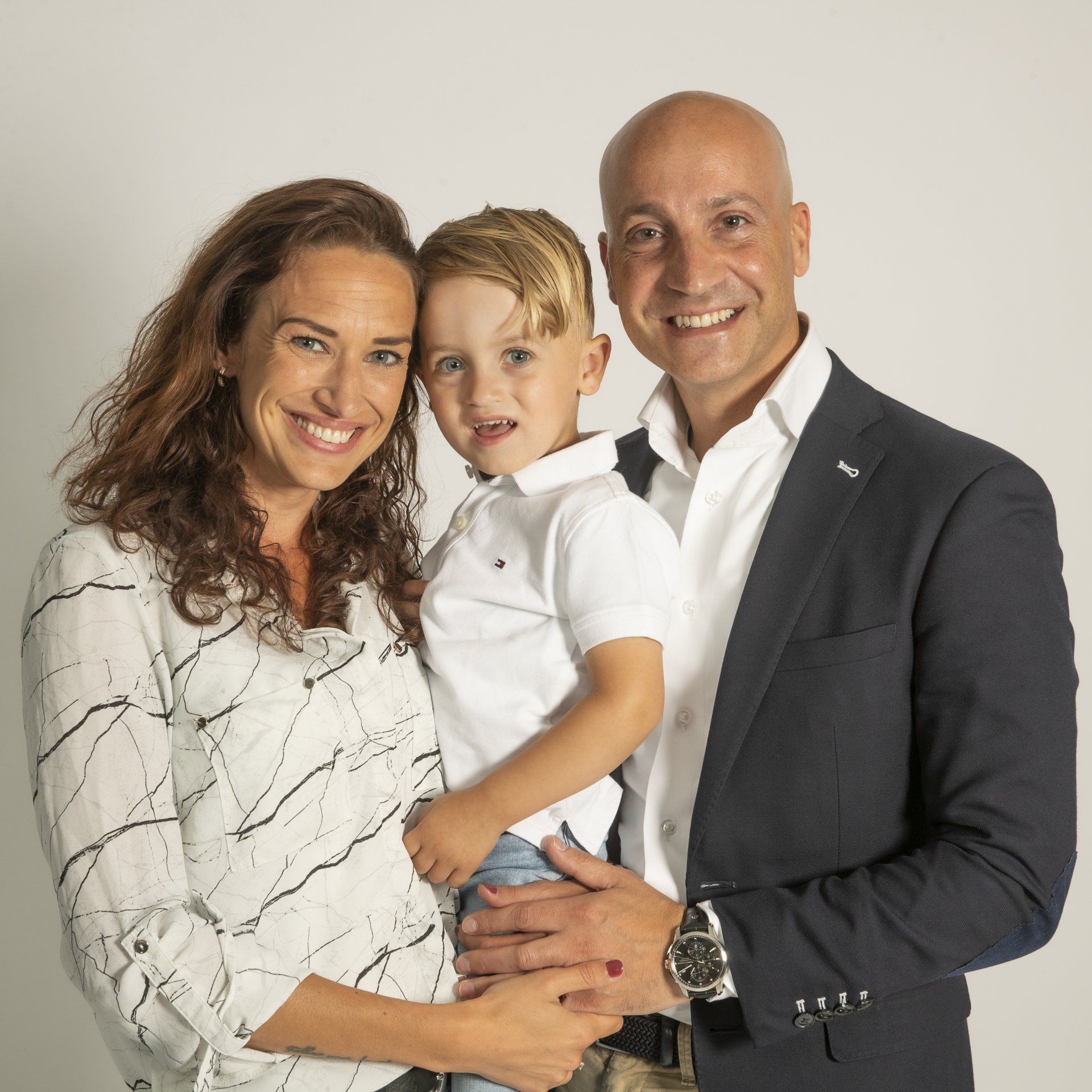 Een lachend stel staat naast elkaar en houdt een jong kind tussen zich in, tegen een effen witte achtergrond.