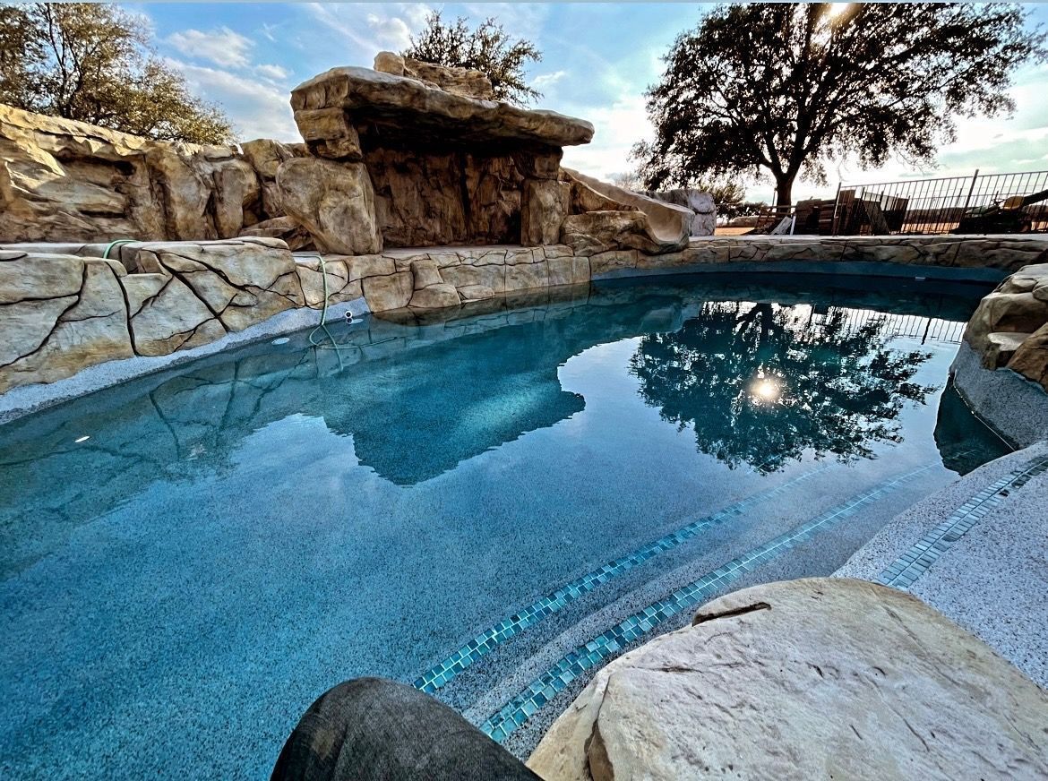 Swimming pool with rocky edges, turquoise water, and a sunlit tree reflected in the water.