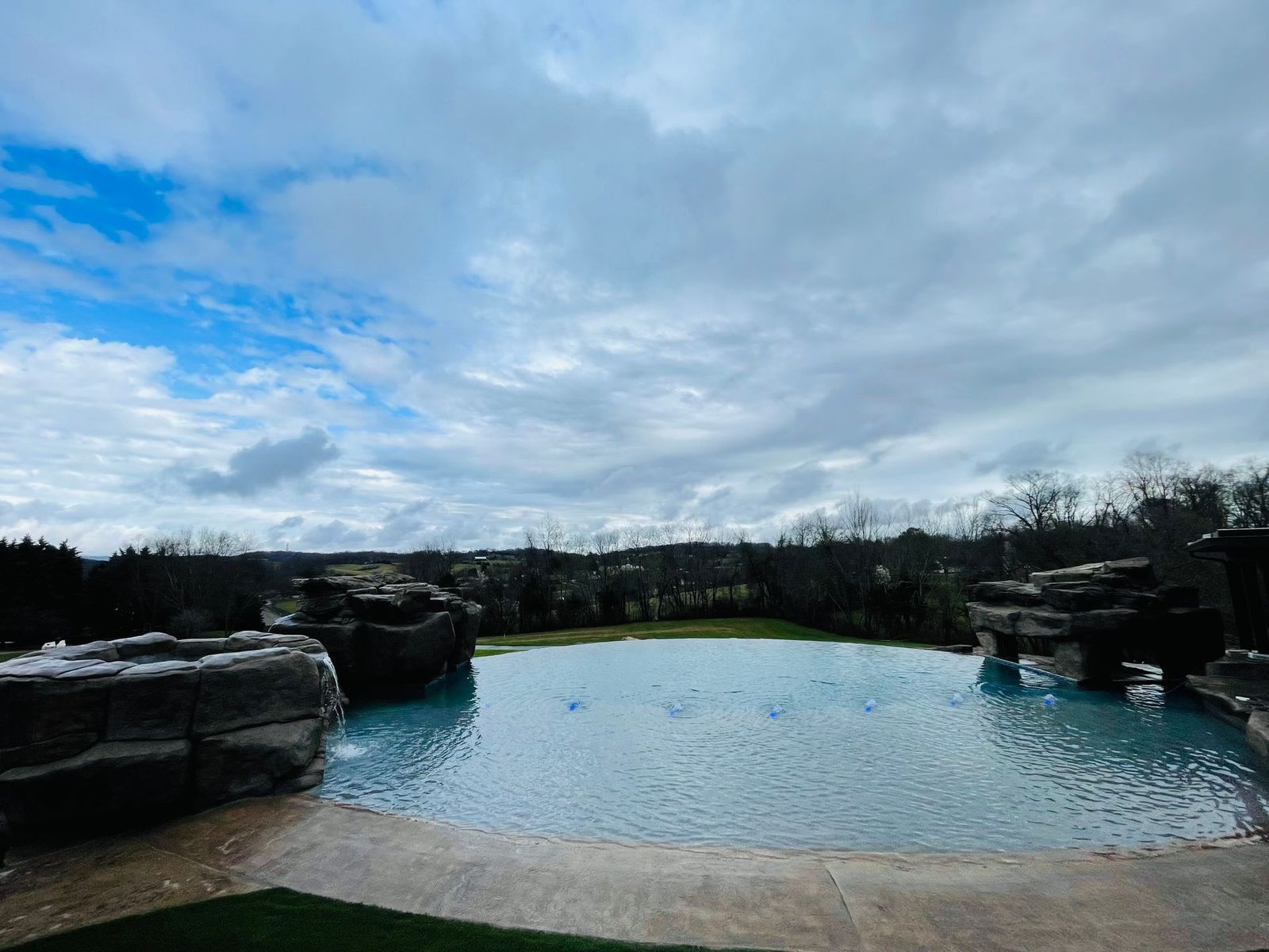 Blue pool surrounded by rocks, under cloudy sky. Trees and a small town are visible in the distance.