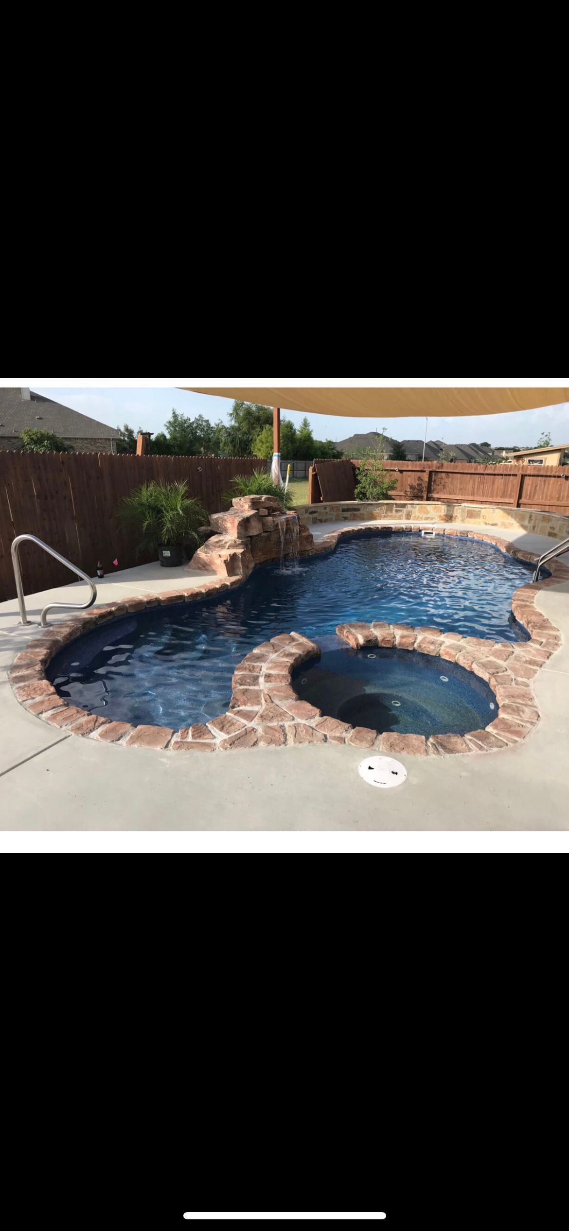 Pool with brick trim, jacuzzi, and rock waterfall. Surrounded by a patio and fence, in an outdoor setting.