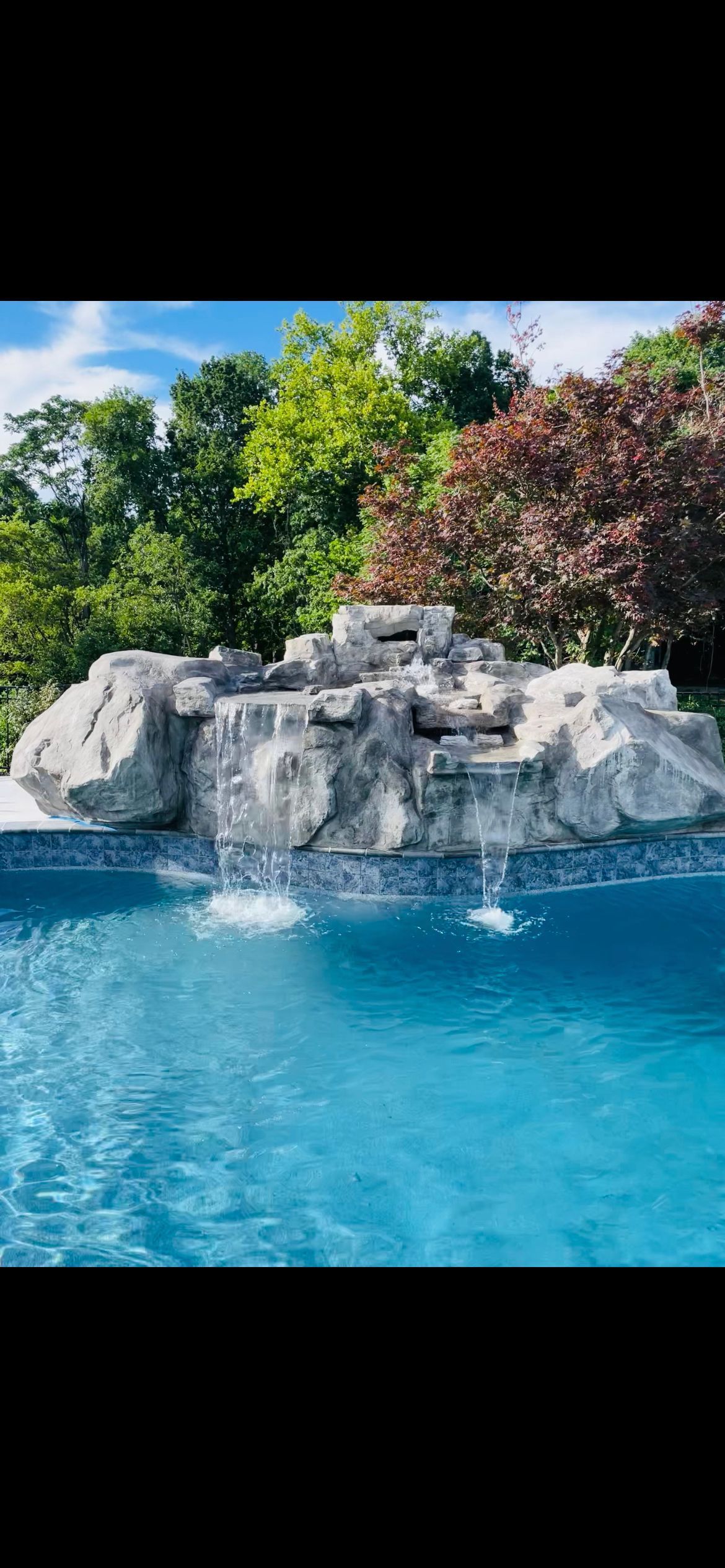 A waterfall cascading into a blue swimming pool, surrounded by rocks and trees.