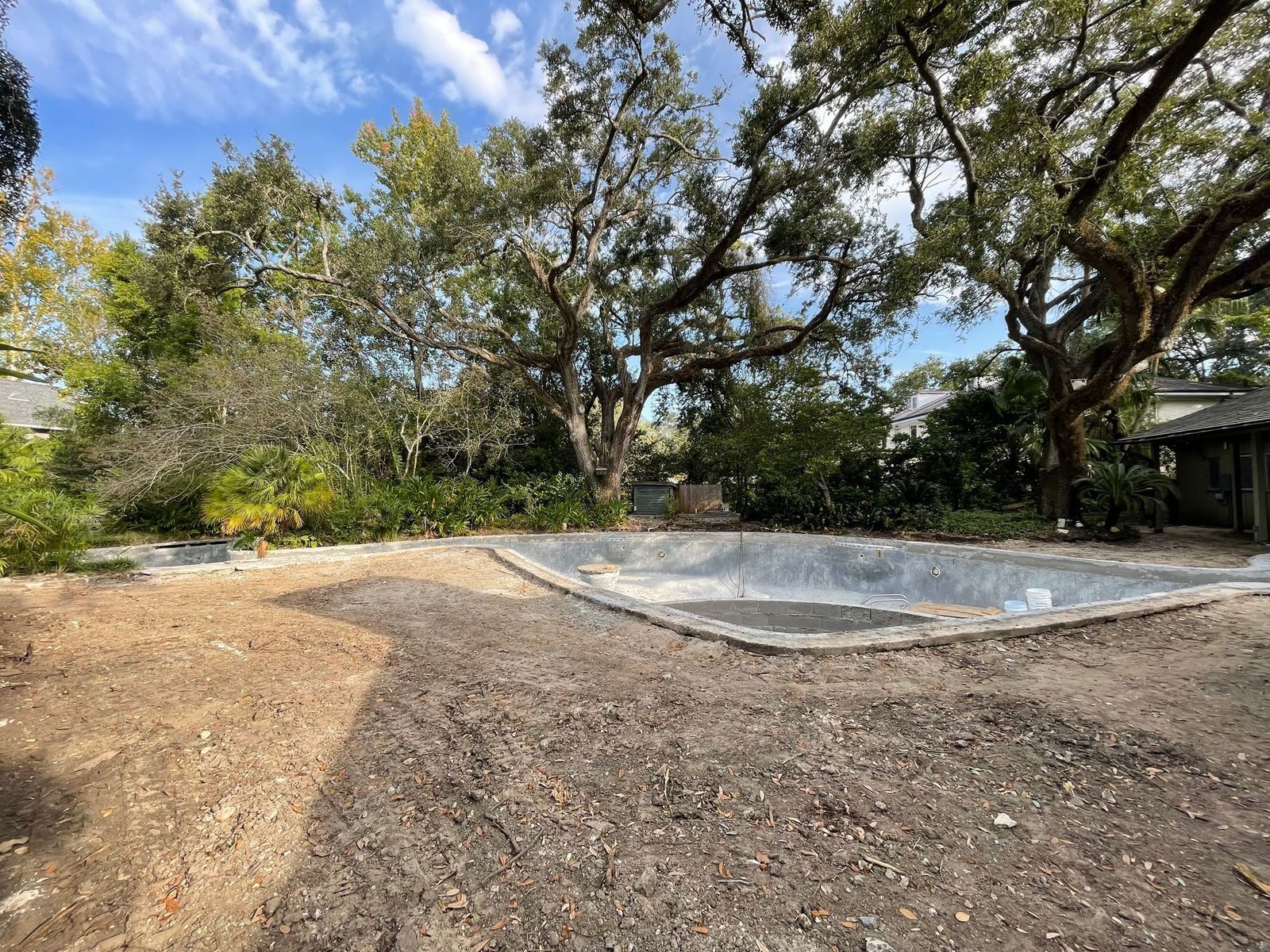 Unfinished backyard with a concrete pool frame, surrounded by dirt and trees. Overcast sky.