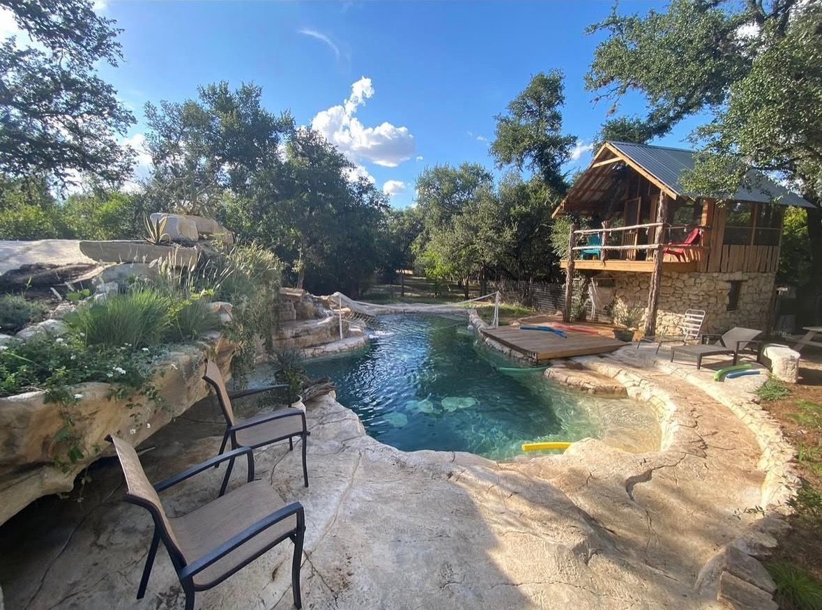 Stone pool with waterfall, two chairs, and a small wooden house with balcony surrounded by trees under a blue sky.