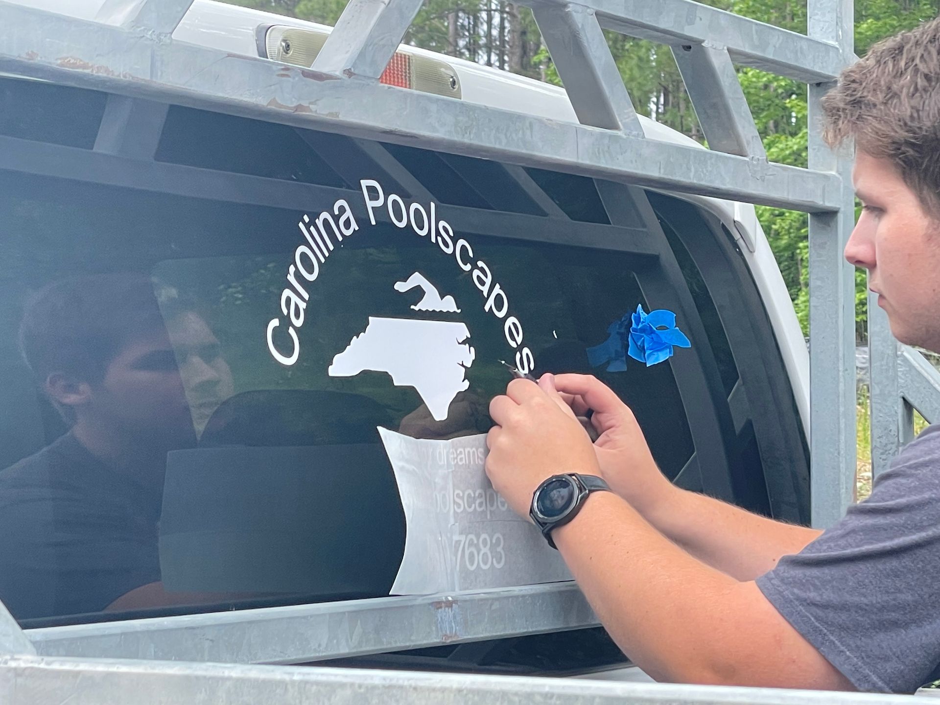 Person applies decal to black vehicle window with Carolina Poolscape logo and North Carolina outline.