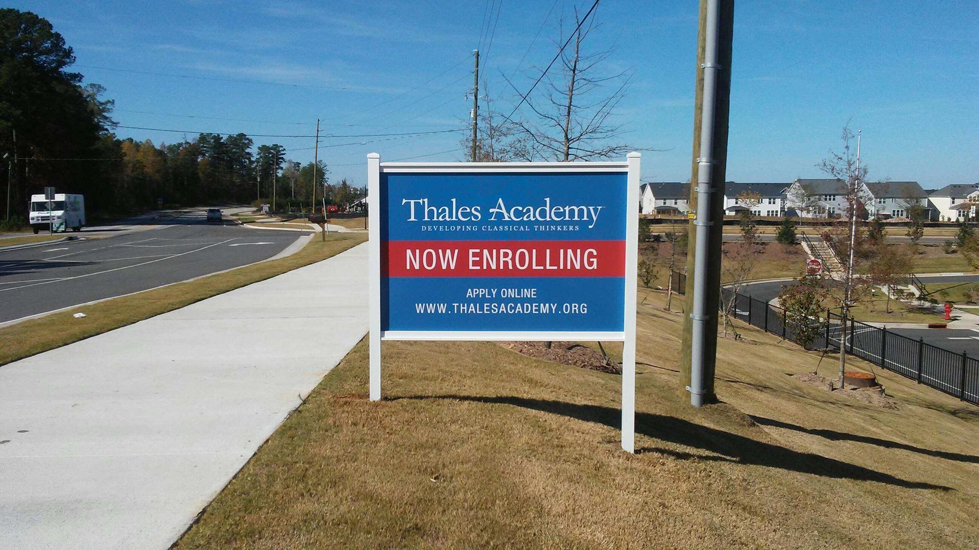 Thales Academy “Now Enrolling” sign beside a sidewalk along a road on a sunny day