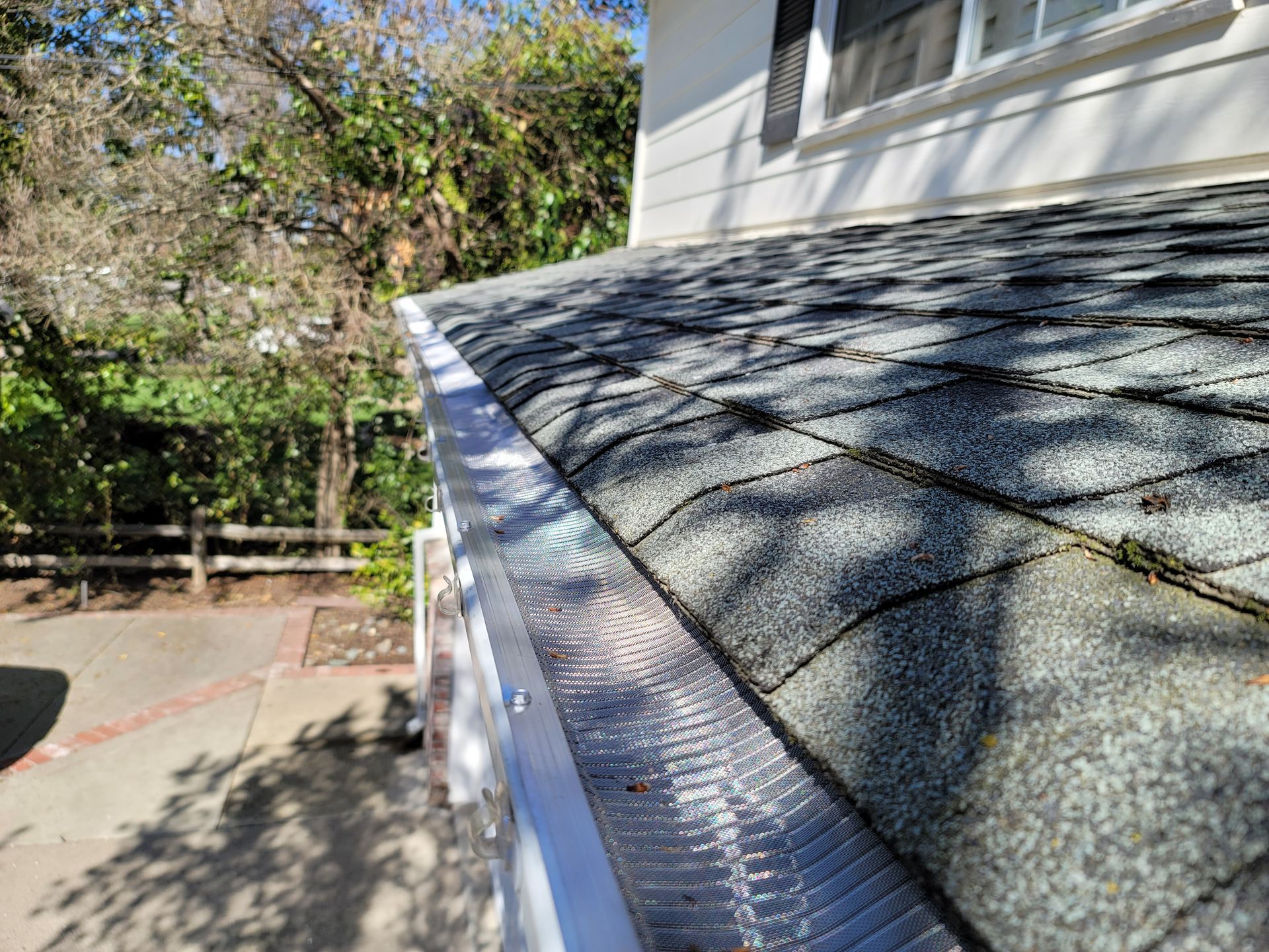 Gutter guard on roof with shingles, near a house. Green trees and foliage are visible in the background.