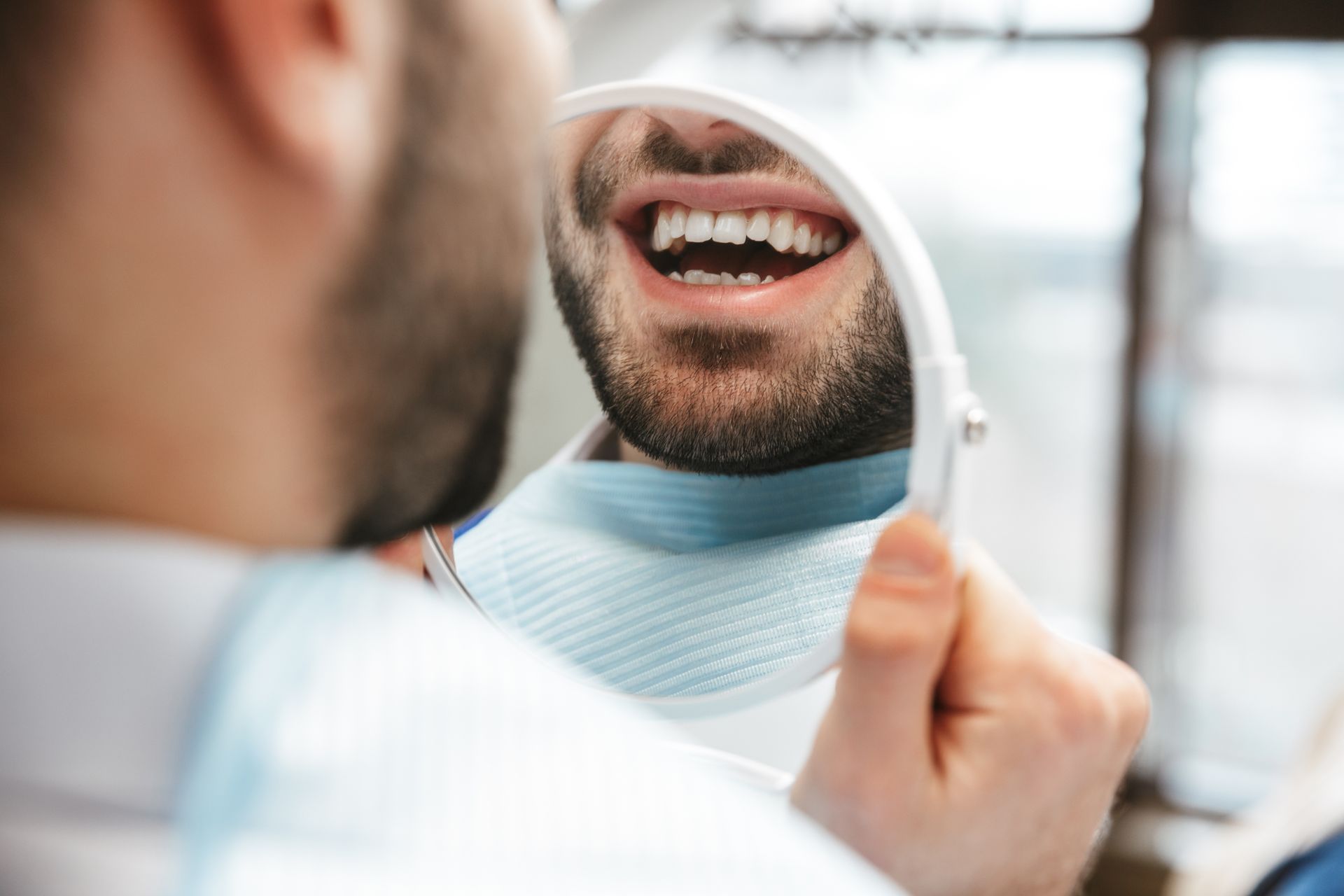 Person examining teeth in a handheld mirror, smiling with an open mouth in a bright room