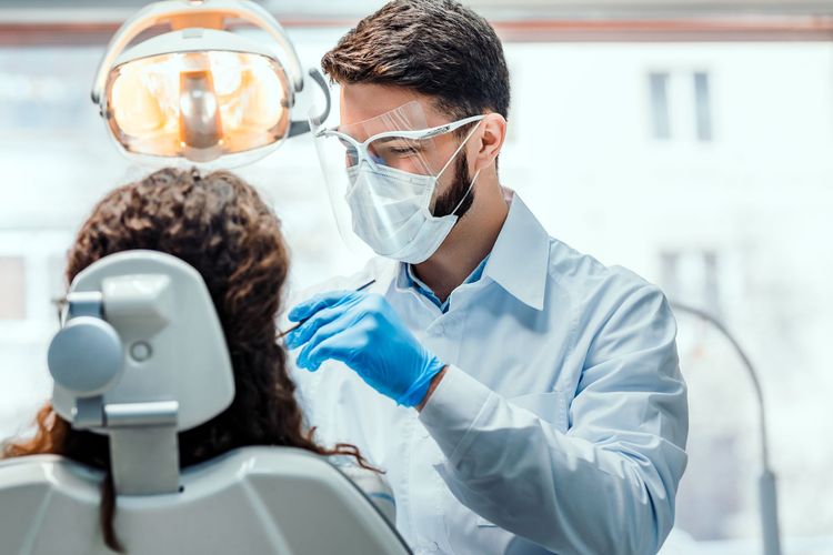 Dentist wearing mask and gloves examines a patient in a dental chair under bright exam light Dentist wearing mask and gloves examines a patient in a dental chair under bright exam light