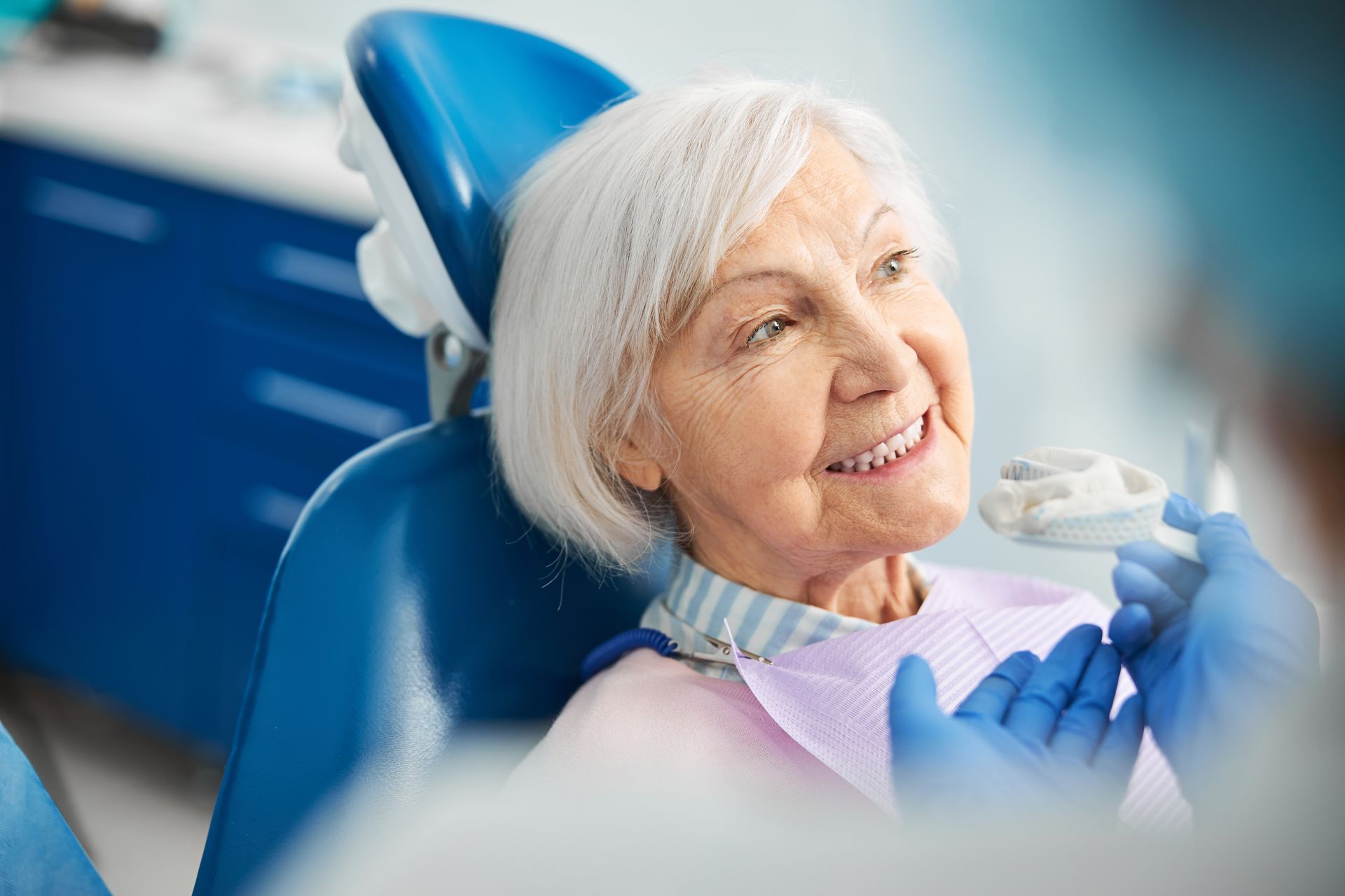 Smiling patient in dental chair during checkup, wearing bib and gloves, with blue clinic background Smiling patient in dental chair during checkup, wearing bib and gloves, with blue clinic background