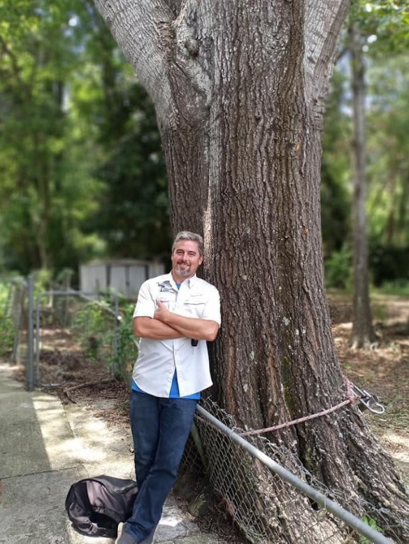 A man is leaning against a tree with his arms crossed.