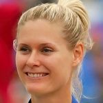 Blonde woman with a ponytail smiles, eyes crinkling. Outdoors, blurred background.