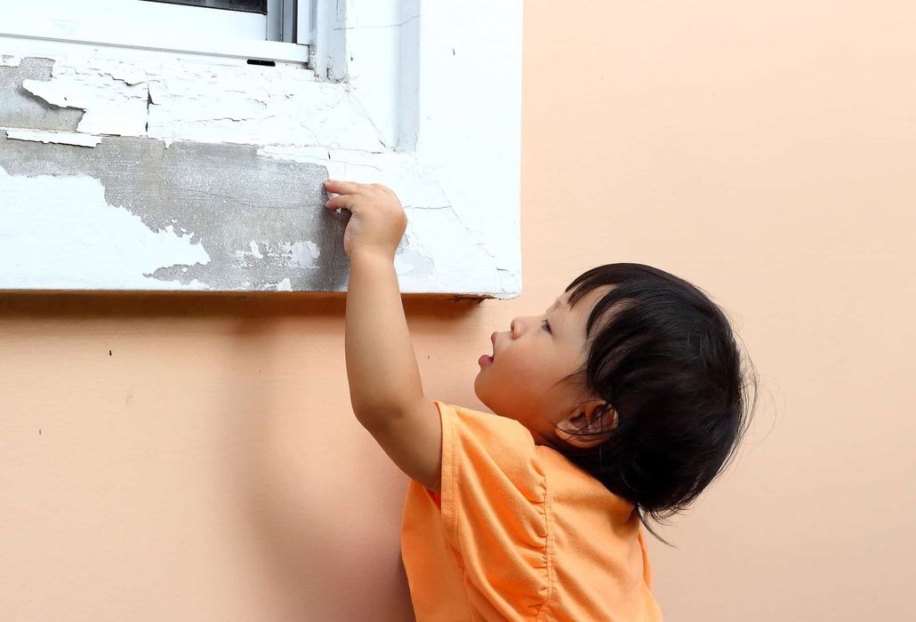 Child in an orange shirt reaching up to touch a peeling white wall outdoors