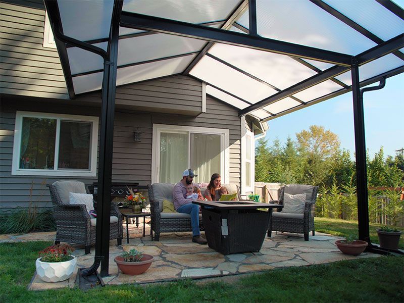 Covered patio with wicker chairs and table where two people sit by a house, overlooking a garden.
