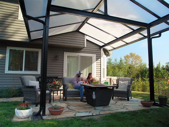 Covered patio with wicker chairs and table where two people sit by a house, overlooking a garden.