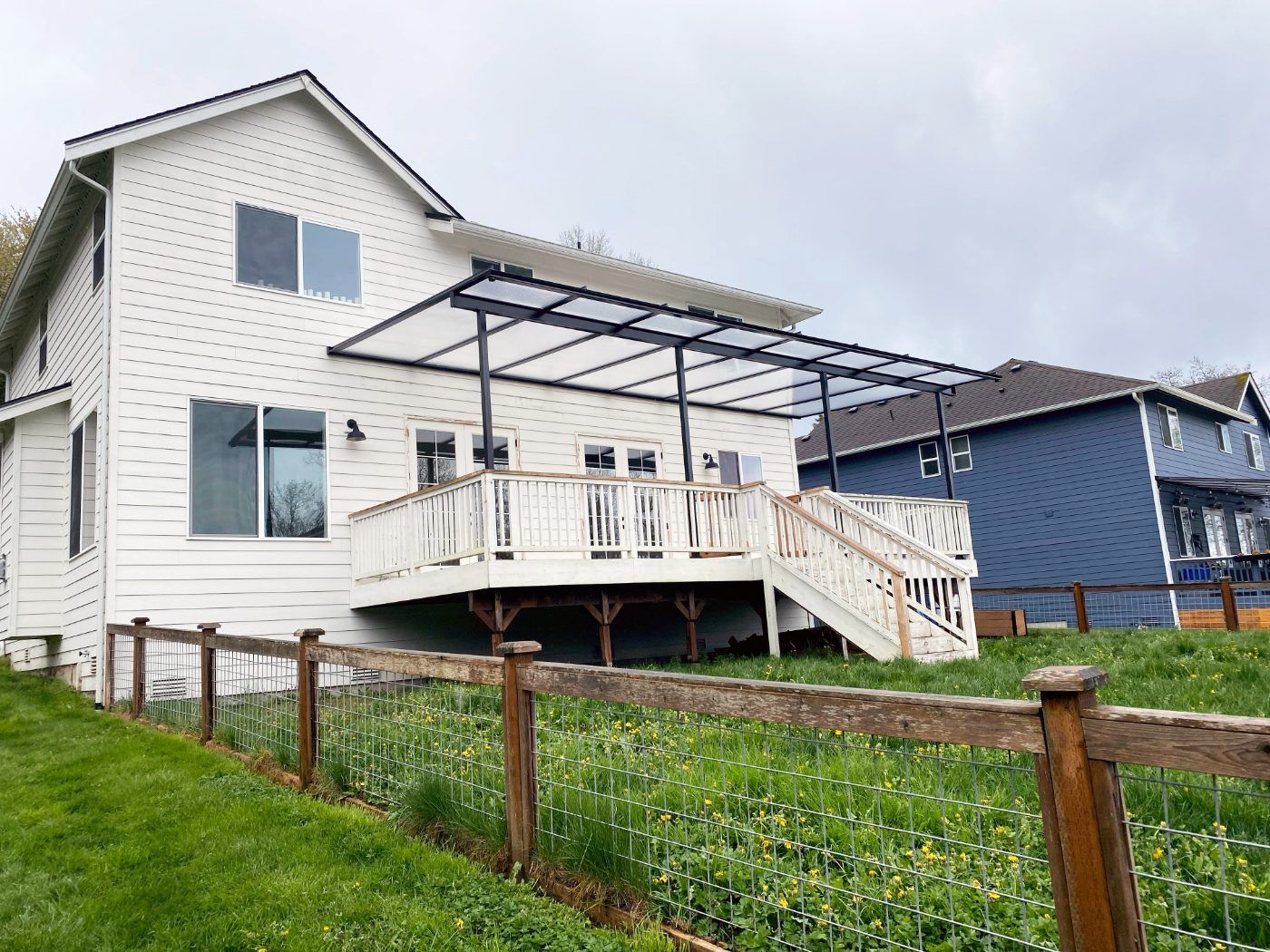 White two-story house with a raised deck, APC patio cover, and stairs on a grassy slope.
