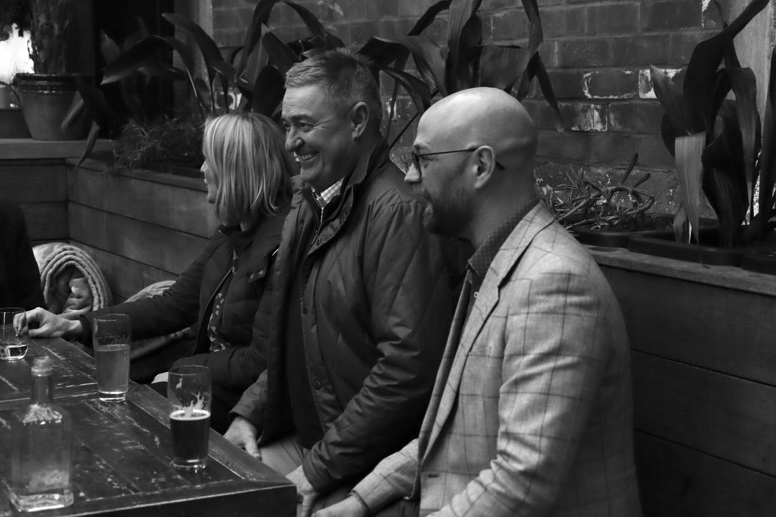 Three people sit at a bar, smiling and talking in a dimly lit room.