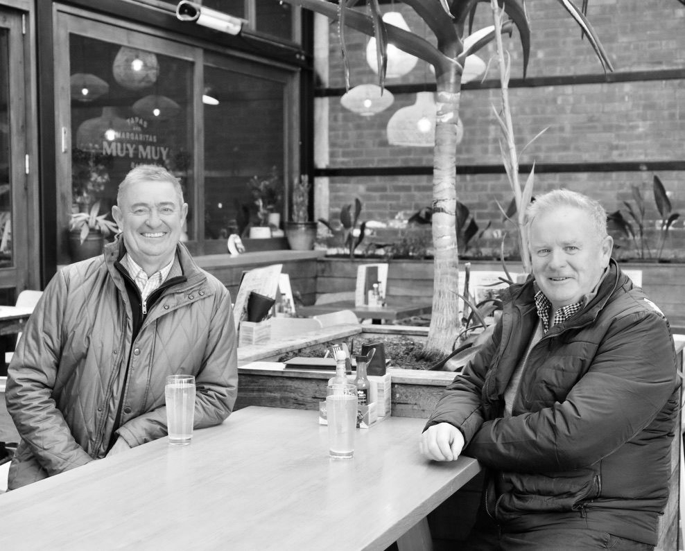 Two people sitting at a café table outdoors, smiling in black-and-white.