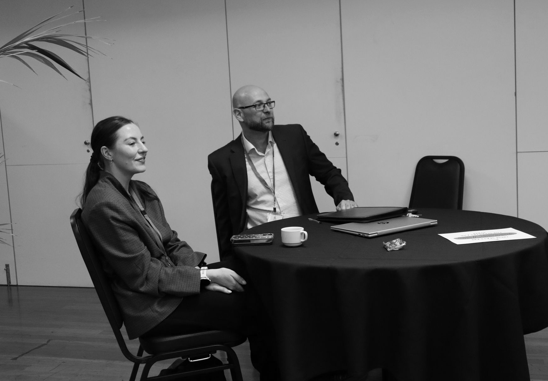 Two people are seated at a round table in a meeting room, looking to the right.
