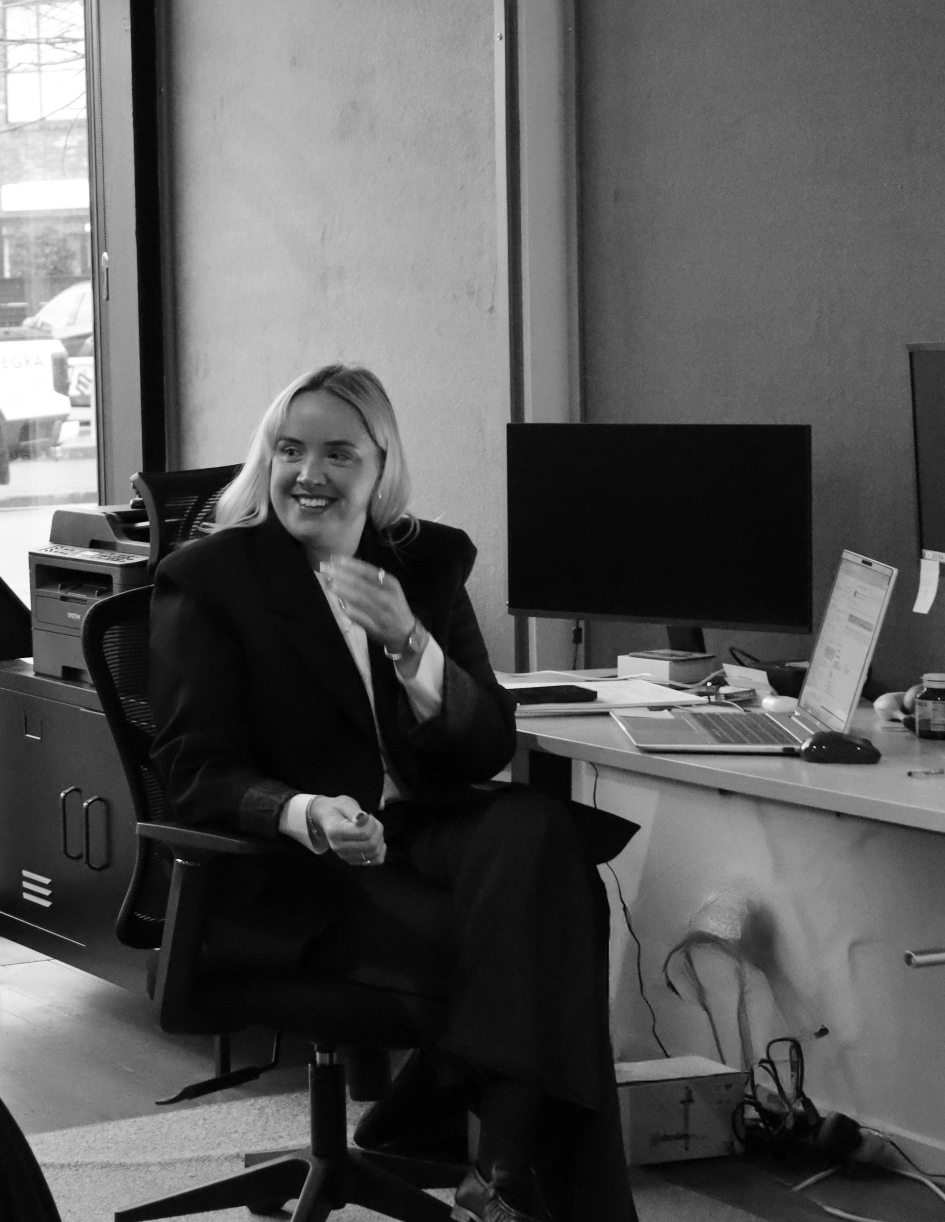 Person sitting in an office chair at a desk, smiling beside a computer monitor.