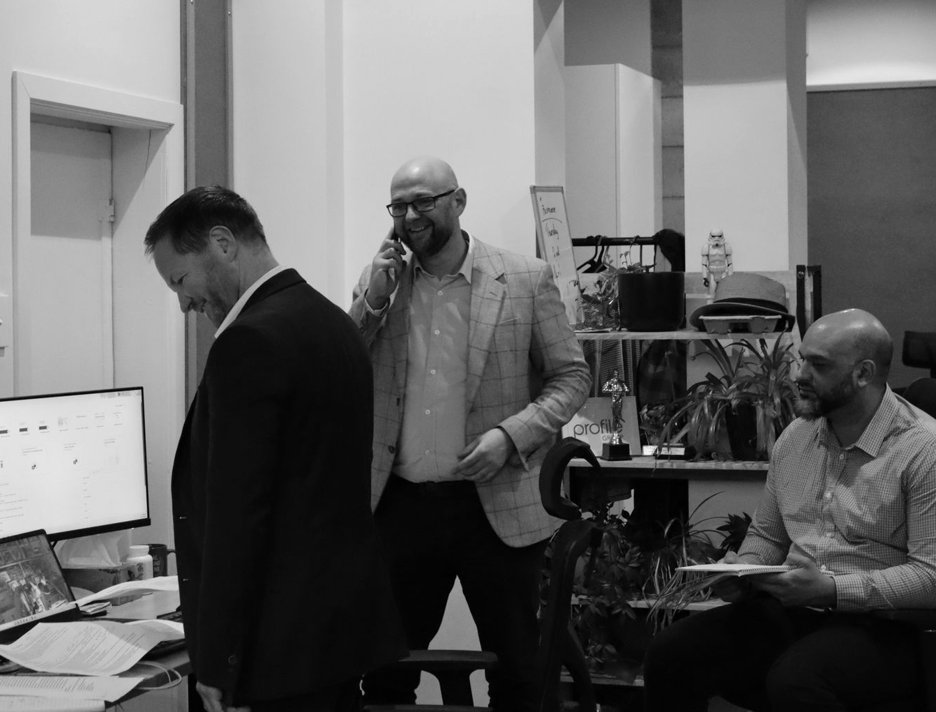 Three people in an office, two standing and one seated near a computer workstation.