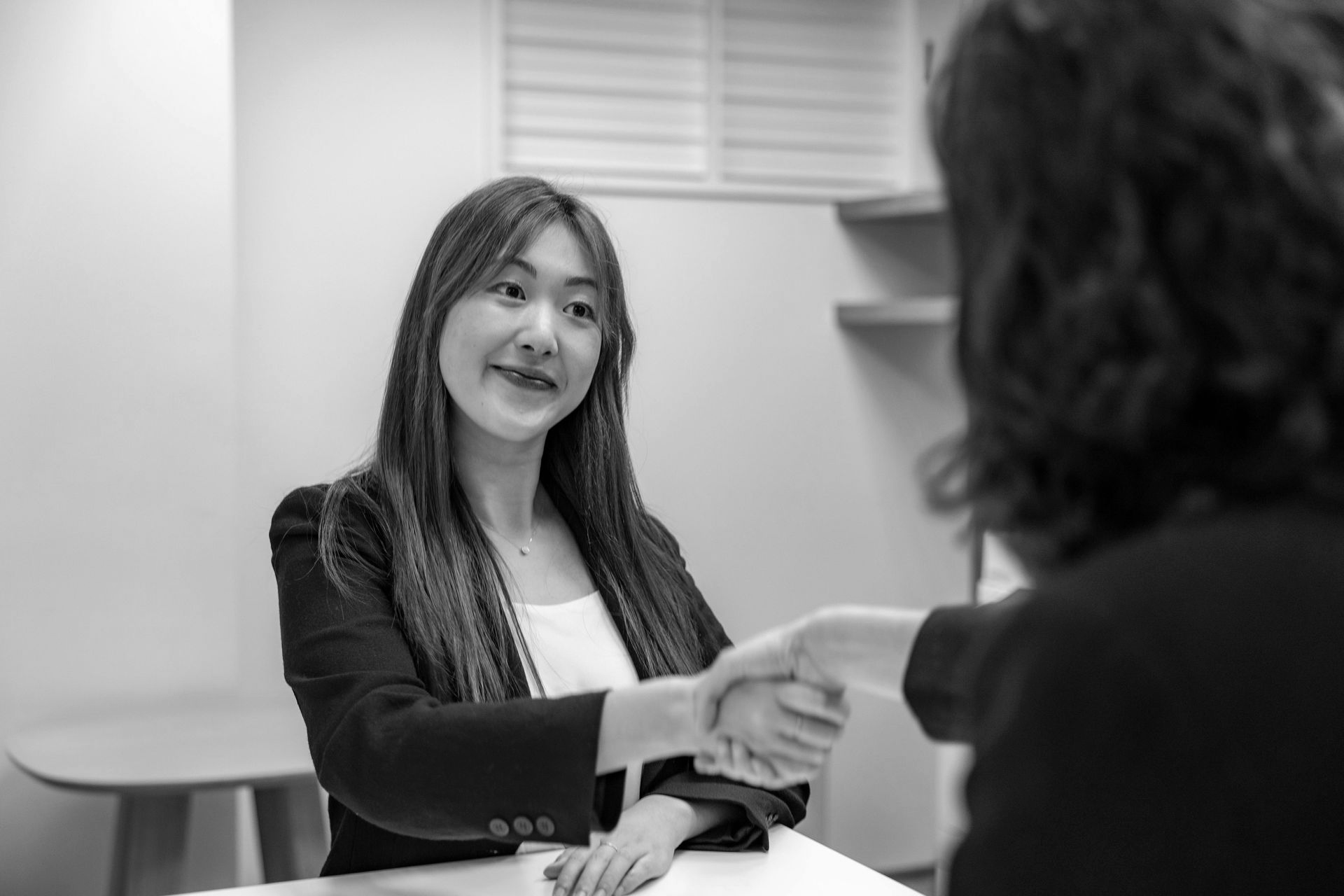 Two people shake hands across a table in a bright office.