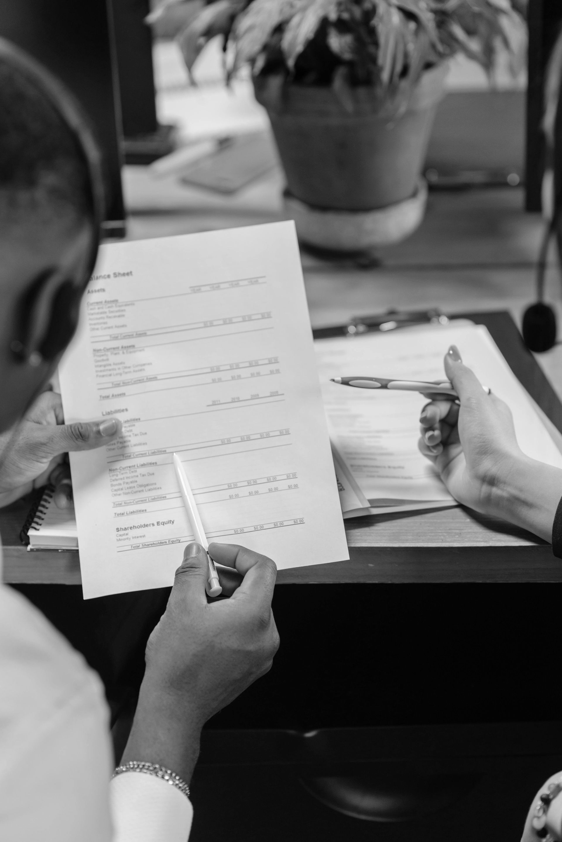Two people reviewing a printed document at a desk with a potted plant in the background