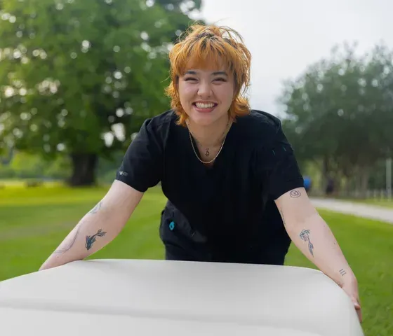 Woman with orange hair smiles, pushing a white bin outdoors, tattoos visible on arms.