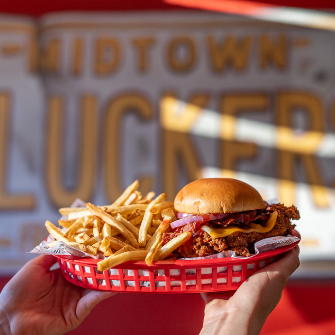 A person holding up a basket of fries with Clucker's Cowboy Clucker Sandwich. In the background is a sign that says