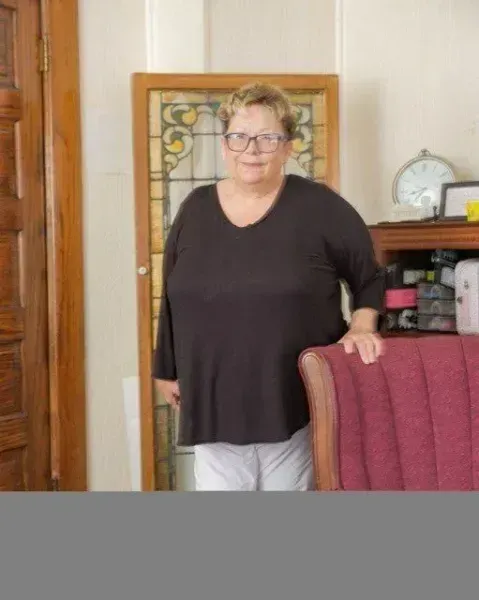 A woman wearing glasses and a black shirt is standing next to a chair in a living room.