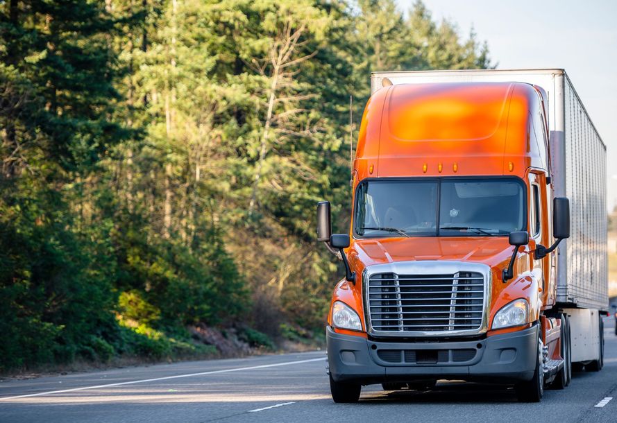 Orange semi-truck driving on a highway, with trees in the background.