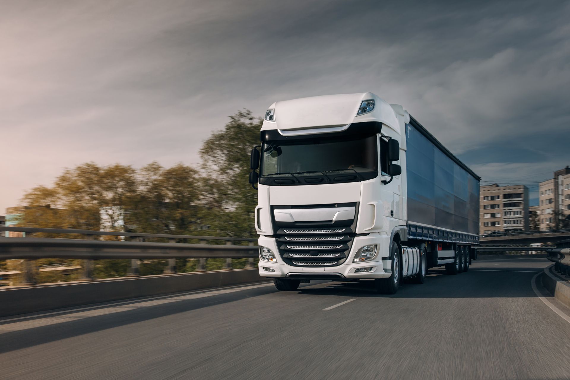 White semi-truck driving on a road with a gray sky and buildings in the background.