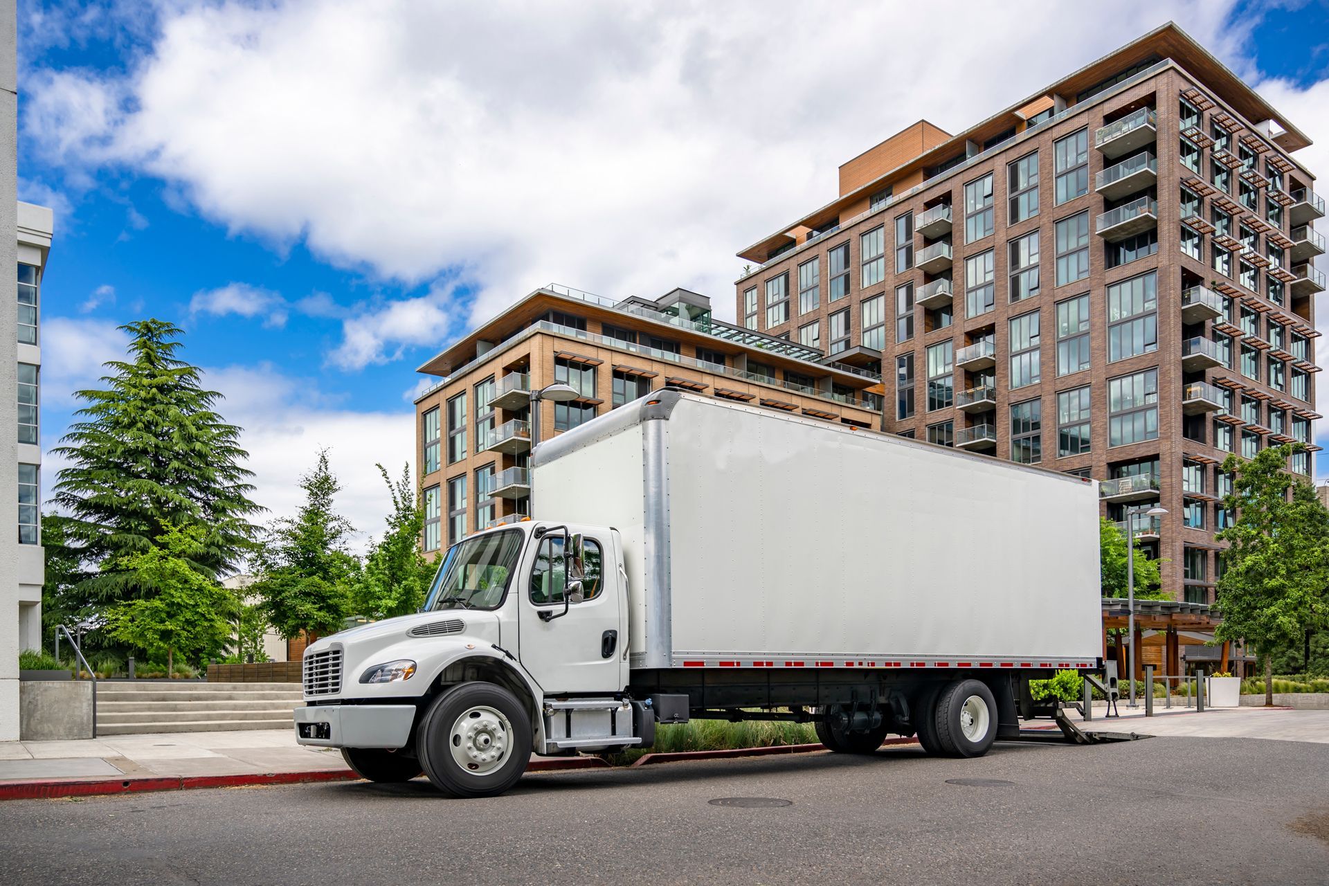White box truck parked on a street in front of a modern apartment building under a cloudy sky.